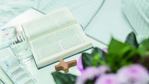 Ein Kreuz und eine aufgeschlagene Bibel liegen neben Medikamenten auf einem Krankenbett-Tisch.